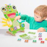 A young child sitting on a play mat, engaged with a wooden Montessori math toy. The toy features a wooden base with numbered slots and colorful finger-shaped counting pieces. The child is holding a small wooden block, focusing on matching it to the corresponding number. Brightly colored stars, circles, and triangles are scattered around the wooden sorting box in a sunlit room.