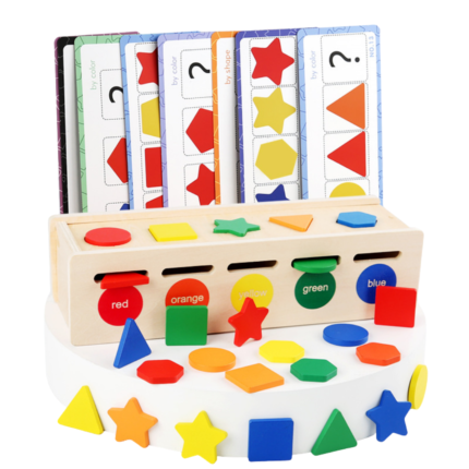 A toddler sitting on a colorful playroom rug, playing with a wooden Montessori-style shape and color sorting toy. The child is wearing blue overalls and a yellow shirt, focused on placing a green wooden square into a slot. Several colorful shapes like stars, circles, and triangles are scattered on the floor around the toy.